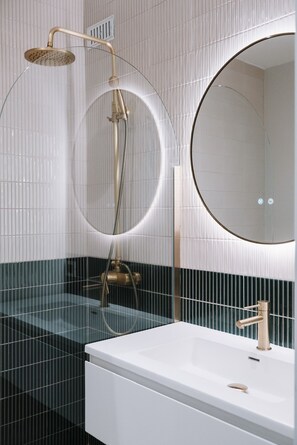 Un baño moderno con un lavamanos blanco, un espejo redondo y una ducha con mampara de cristal curva.