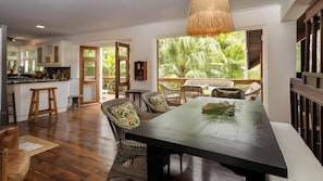 A spacious dining area with a long table and wicker chairs, a kitchen with white cabinets, and a large window overlooking a tropical garden. - Dragonfly Villa - By Zenbreak (Holetown)