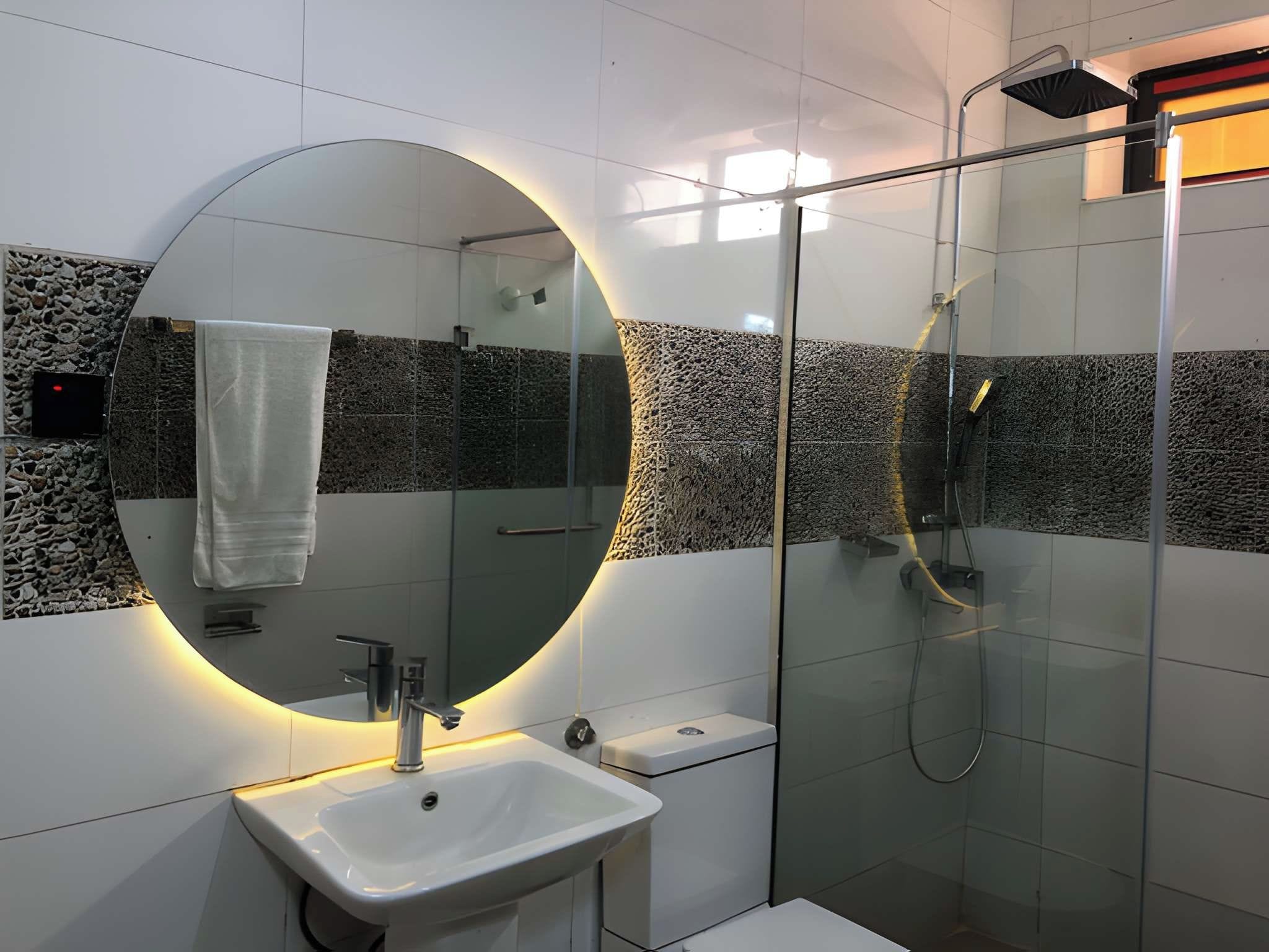 Un baño moderno con un espejo redondo, un lavamanos y una ducha con mampara de vidrio.