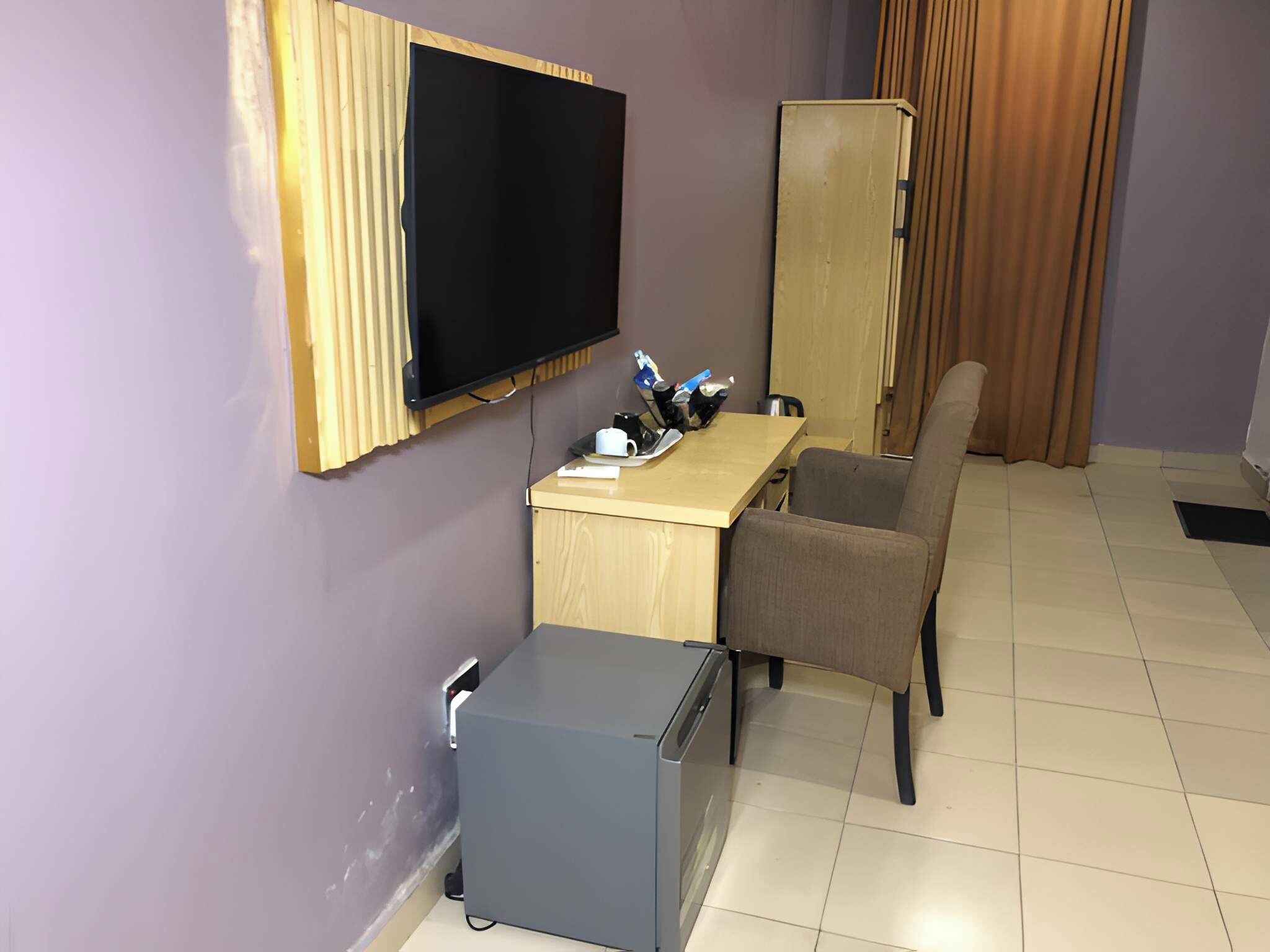 A compact room with a flat-screen TV mounted on the wall, a small refrigerator, a desk with a chair, and a cabinet.