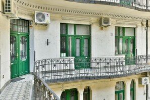 A building with green doors and windows, a balcony with a wrought iron railing, and air conditioning units on the exterior. -  King of Church Budapest- Church-View Balcony Apartment  in Central Budapest (Budapest)