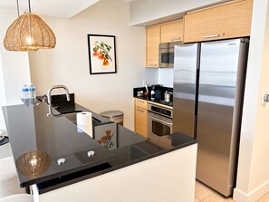 A modern kitchen with a stainless steel refrigerator, a microwave, and an oven. There is a black countertop with a sink and a wicker light fixture. - 24th Floor 1-bedroom Full Kitchen Washer/Dryer Sunny Isles Direct Beach Access (Sunny Isles Beach)