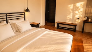 Una cama bien hecha con cabecera, una mesita de noche con lámpara, y un banco en una habitación con suelo de madera y una puerta al fondo.