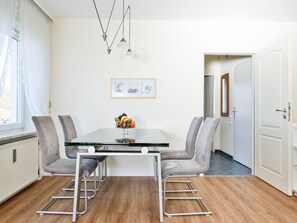 Ein Essbereich mit einem Glastisch, vier grauen Stühlen, einer Obstschale, Pendelleuchten und einem gerahmten Bild an der Wand.