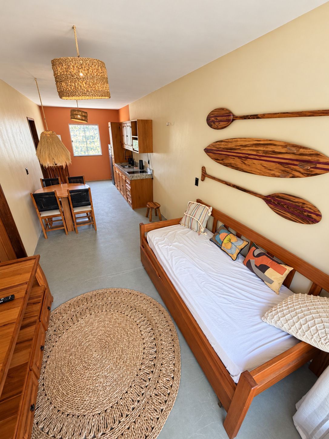 Ein Schlafzimmer mit einem hölzernen Bett, einem gewebten Teppich, einer Essecke mit Tisch und Stühlen sowie einer Küche mit einer Pendelleuchte.