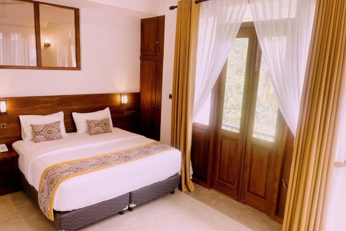 A hotel room with a large bed, wooden headboard, a nightstand, a mirror, and a door with curtains.
