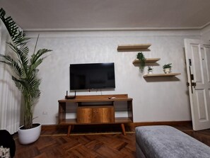 A living room with a flat-screen TV mounted on the wall, a wooden entertainment unit below, and three wooden shelves with plants. - New Listing (Miraflores)