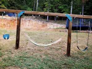 A hammock between two wooden posts in a grassy area with a wooden swing set and a blue structure in the background. - Brook Highland House (Birmingham)
