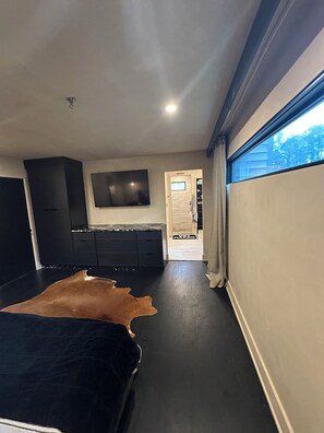 A modern bedroom with a large bed, a flat-screen TV mounted on the wall, and a view of the outdoors through a window. - Tiki Oasis at Indian Circle (Kennesaw)
