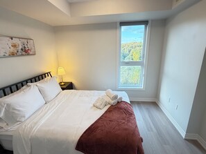 A bedroom with a bed, a nightstand, a lamp, and a window with a view of trees. - Scenic Kitchener Retreat | Escarpment View Balcony (Kitchener)