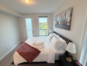 Quarto com cama, mesa de cabeceira, luminária, pintura e vista através de uma porta.