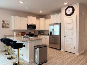 A modern kitchen with white cabinets, a stainless steel refrigerator, a microwave, and a built-in oven. There are bar stools and a trash bin. - Brand-New home 5 Min to downtown, near beaches, community pool (Port St. Joe)