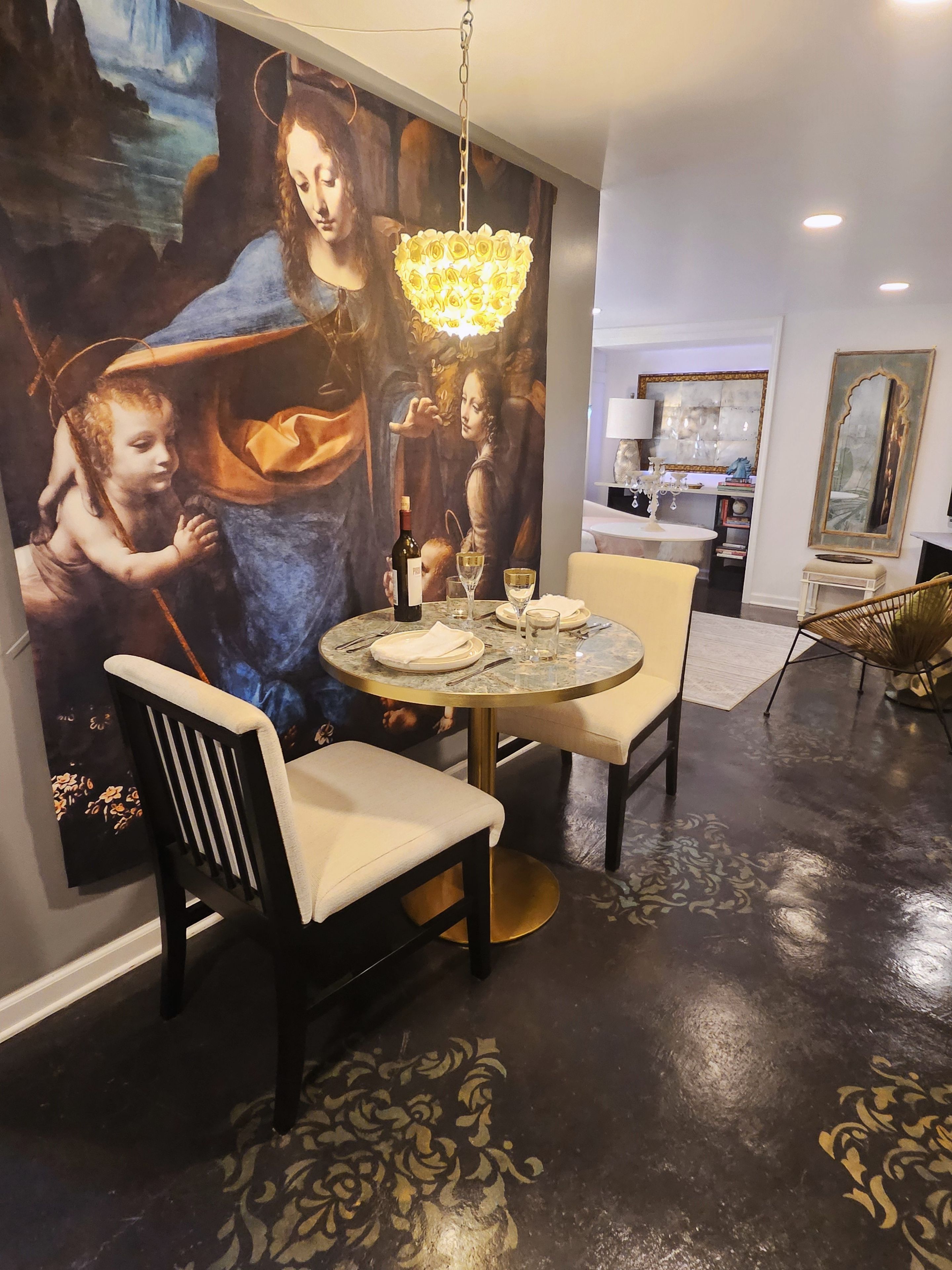 A dining area with a round table, chairs, a chandelier, and a large painting on the wall.