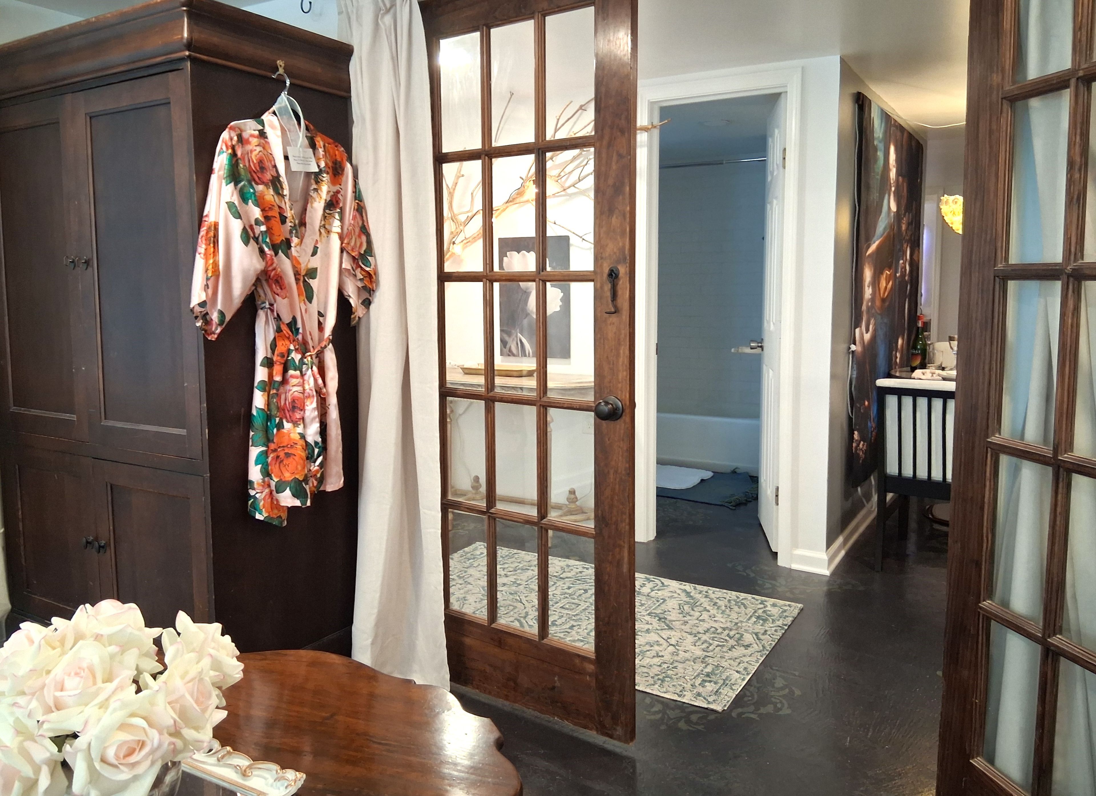 A room with a wooden wardrobe, a floral robe hanging, a vase of flowers, and a view into a bathroom with a bathtub.