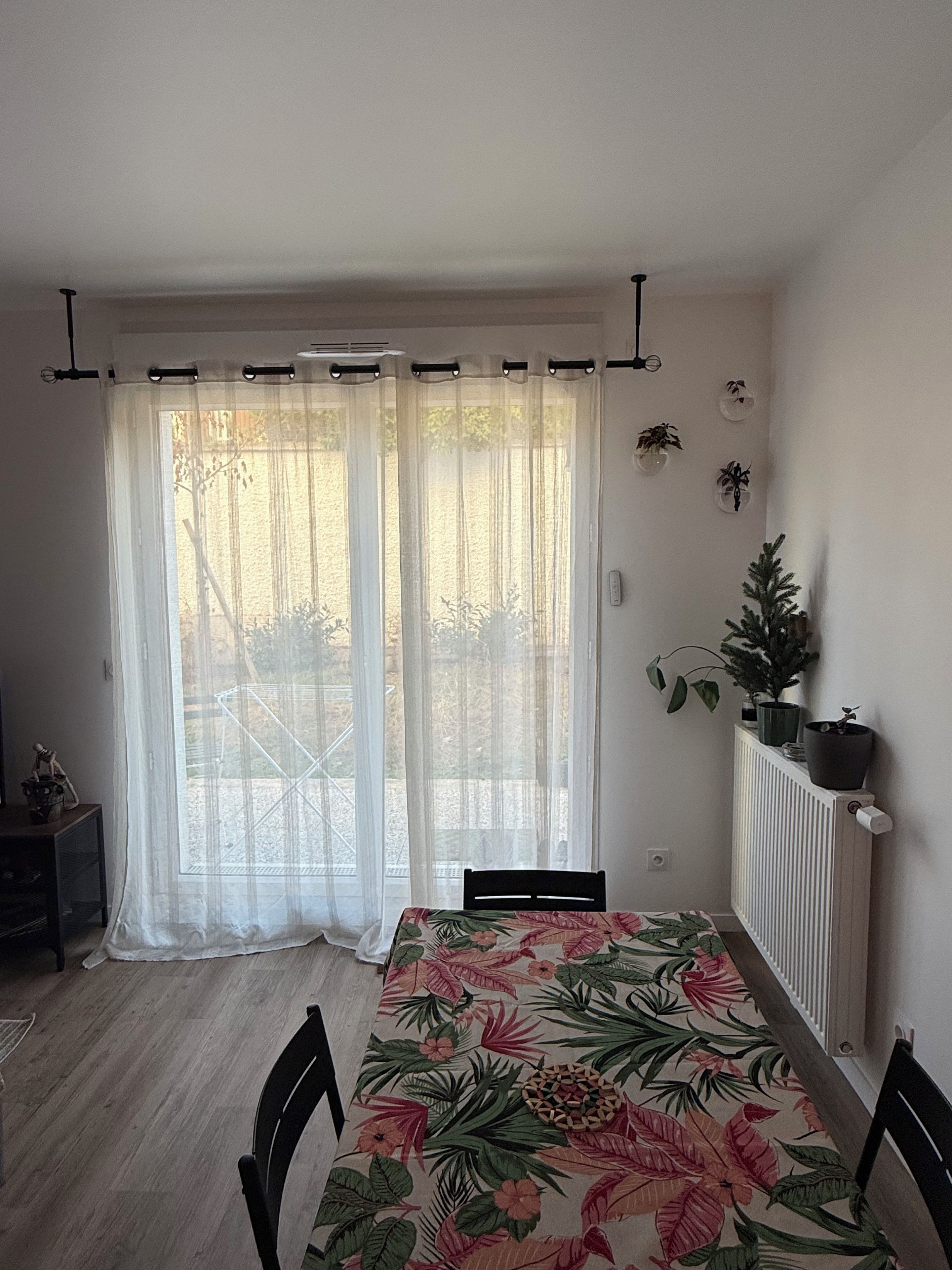 Ruokailutila, jossa on pöytä ja tuolit, ikkuna, jossa on läpikuultavat verhot, patteri ja pieni kasvi patterin päällä.