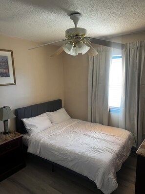 Un dormitorio con cama, mesita de noche, lámpara, ventilador de techo y ventana con cortinas.