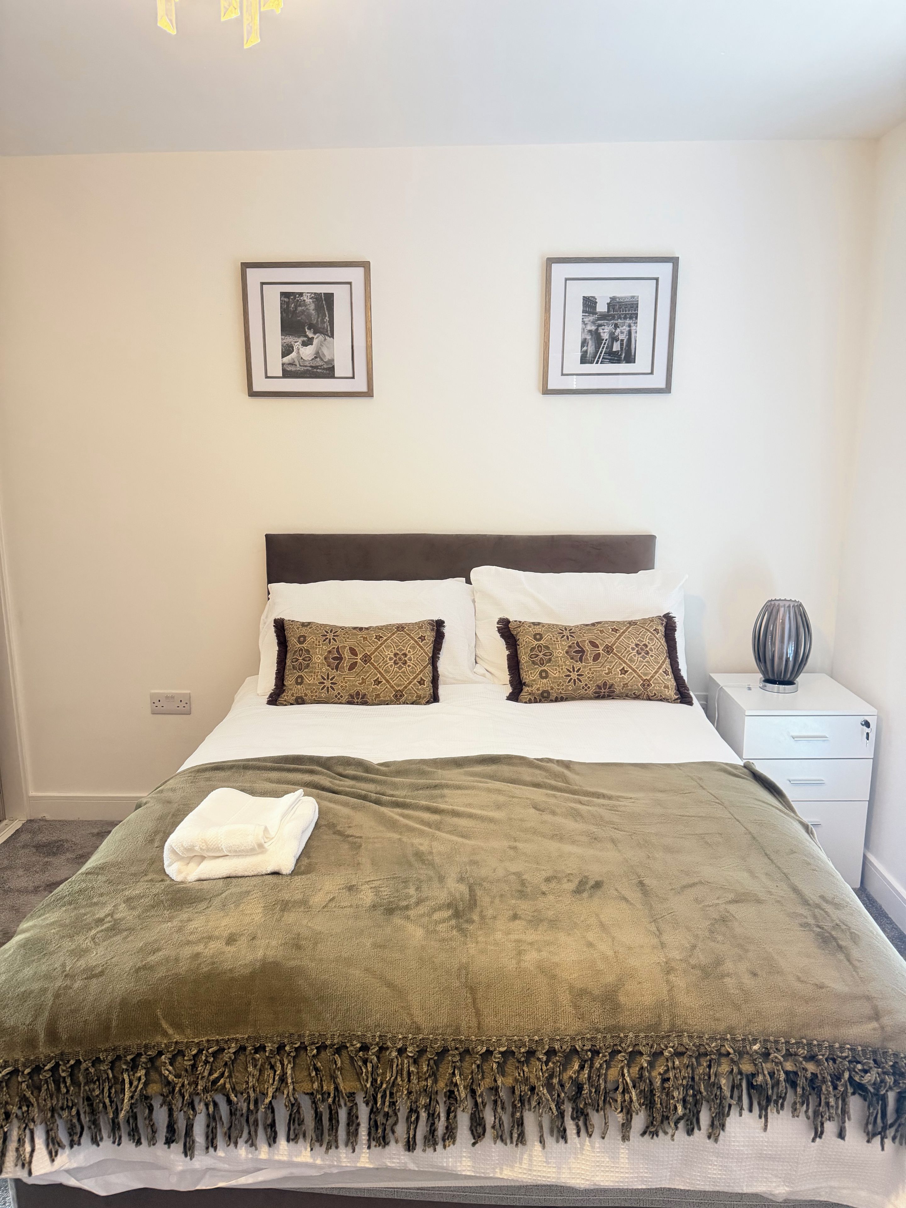 A neatly made bed with a green blanket and patterned pillows, a bedside lamp, and two framed pictures on the wall.