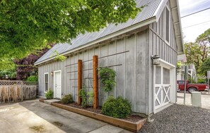 Ein einstöckiges Gebäude mit grauer Holzfassade, einer weißen Tür und einem kleinen Garten mit Pflanzen und einem hölzernen Spalier.