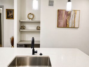 A kitchen with a stainless steel sink, a black faucet, and a white countertop. There are decorative items on the shelves and a painting on the wall. - Chic & Homey Apartment 2BR/1BA Austin TX (Austin)