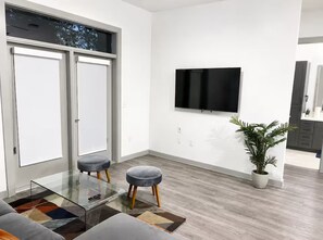 Ein modernes Wohnzimmer mit einem Glastisch, zwei grauen Hocker, einer Topfpflanze und einem flachbildfernseher an der Wand.