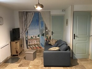A living room with a sofa, a television on a wooden cabinet, a small ottoman, and a view through a window with curtains. - New Listing (Romans-sur-Isère)