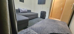A room with a grey sofa, a small table, a suitcase, and a wooden door. - New Listing (Arrahma)