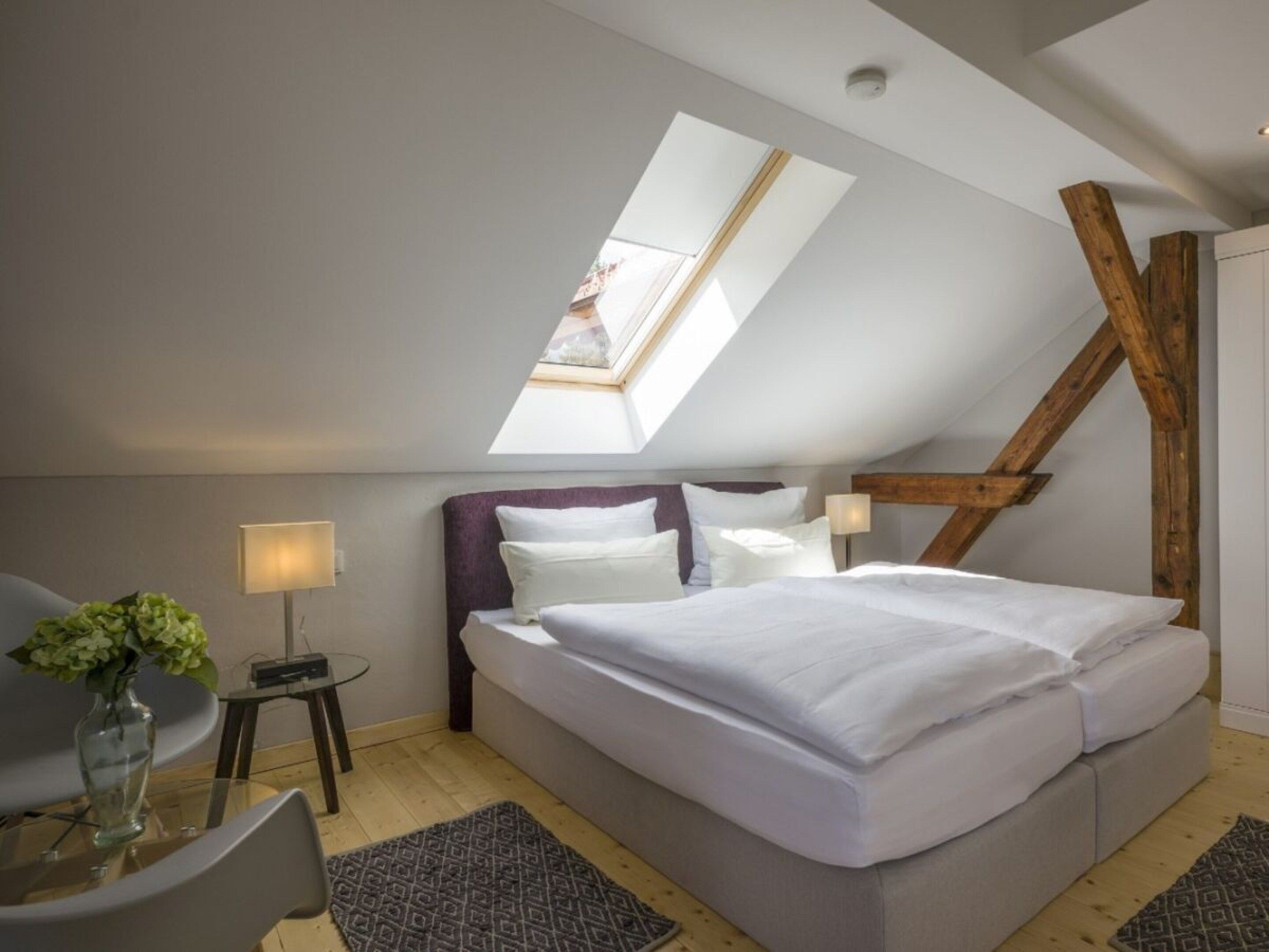 A bedroom with a sloped ceiling, a bed, a chair, a small table, and a vase with flowers.
