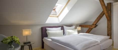 A bedroom with a sloped ceiling, a bed, a chair, a small table, and a vase with flowers.