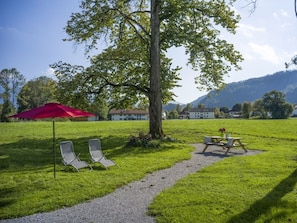 A grassy outdoor area with a picnic table and two lounge chairs under a red umbrella, with a tree and buildings in the background. - Holiday Apartment With 1 Bedroom (Oberaudorf)