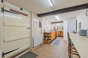 A kitchen with a large white door, wooden cabinets, a radiator, and a dining table with chairs. - Hot Tub & Games Room | Sleeps 11 | 4BR Wyboston (Wyboston)