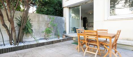 A patio with a wooden table and chairs, a concrete floor, and a stone wall with plants.