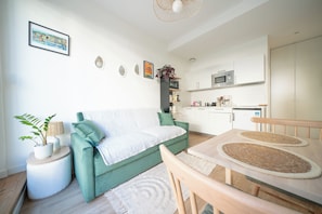 Ein modernes Wohnzimmer mit einem grünen Sofa, einem Esstisch mit gewebten Untersetzern, einer Küchenzeile mit weißen Schränken und einem kleinen Kühlschrank.