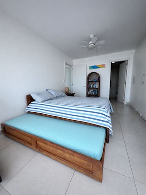 Un dormitorio con una cama individual, un ventilador de techo, una estantería y una puerta que da a otra habitación.