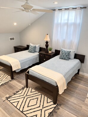 A bedroom with two beds, a ceiling fan, a nightstand with a lamp, and a window with curtains. - Serene 2-bedroom house with AC in Bodden Town (Bodden Town)