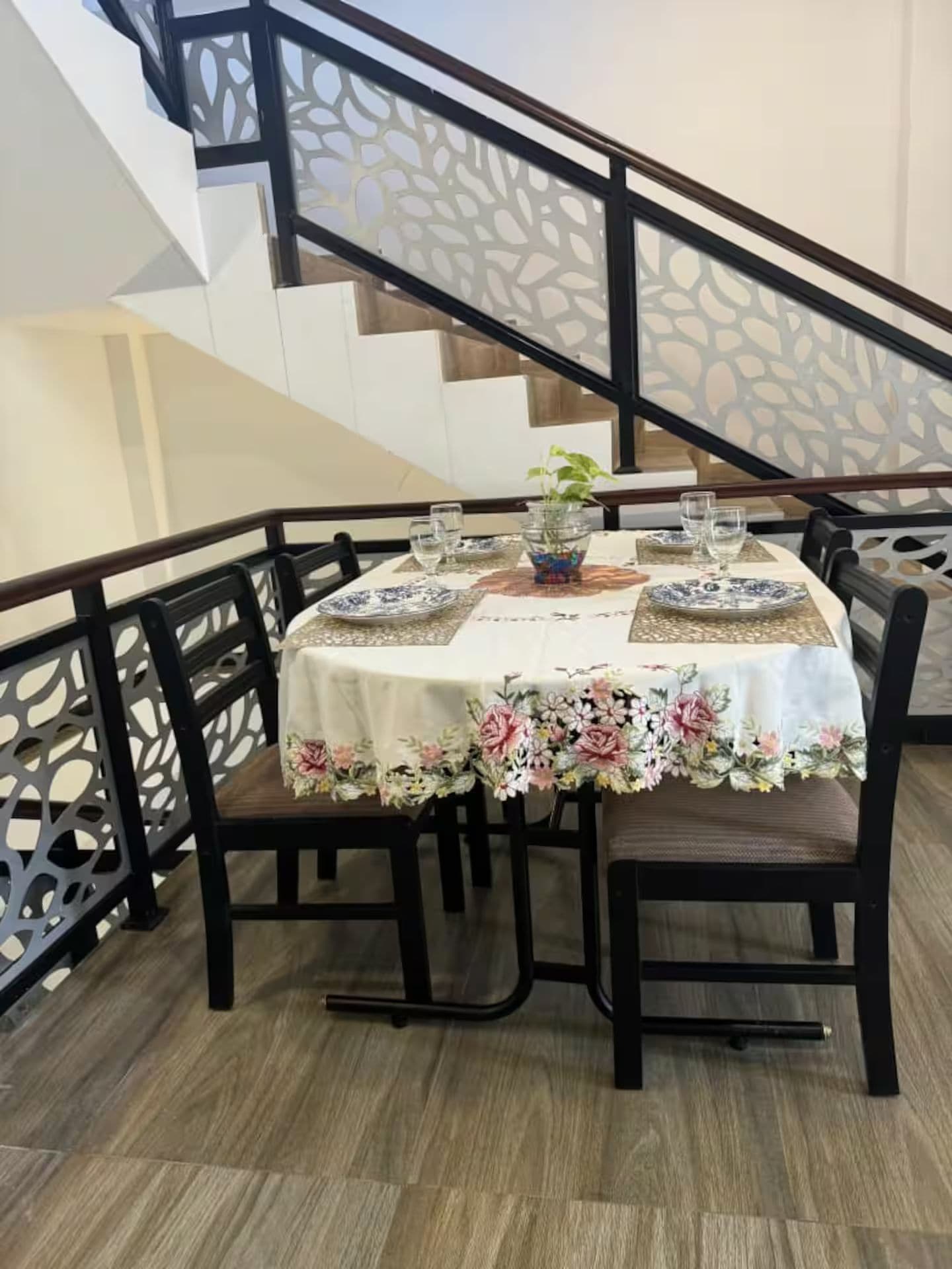 A dining table set for four with floral tablecloth, surrounded by chairs, in a room with a staircase and decorative railing.