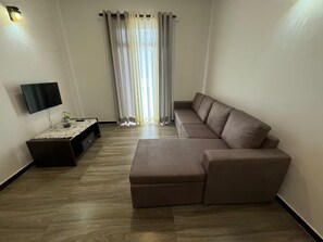 Een woonkamer met een hoekbank, een flatscreen televisie aan de muur, een bijzettafeltje met een plant en een raam met gordijnen.