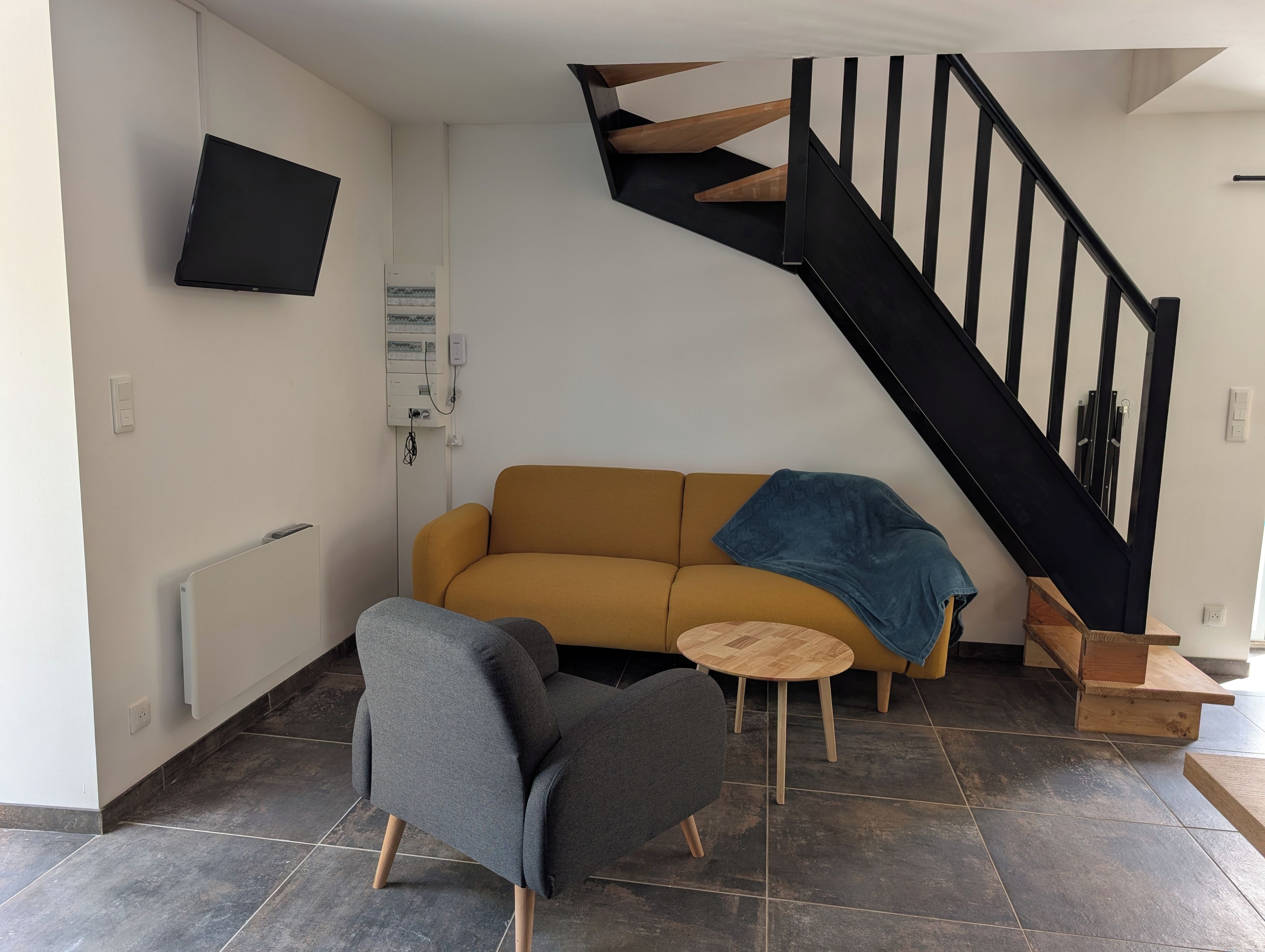 Ein Wohnzimmer mit einer Couch, einem Sessel, einem kleinen runden Tisch, einem an der Wand befestigten Fernseher und einer Treppe mit Holzhäuschen.