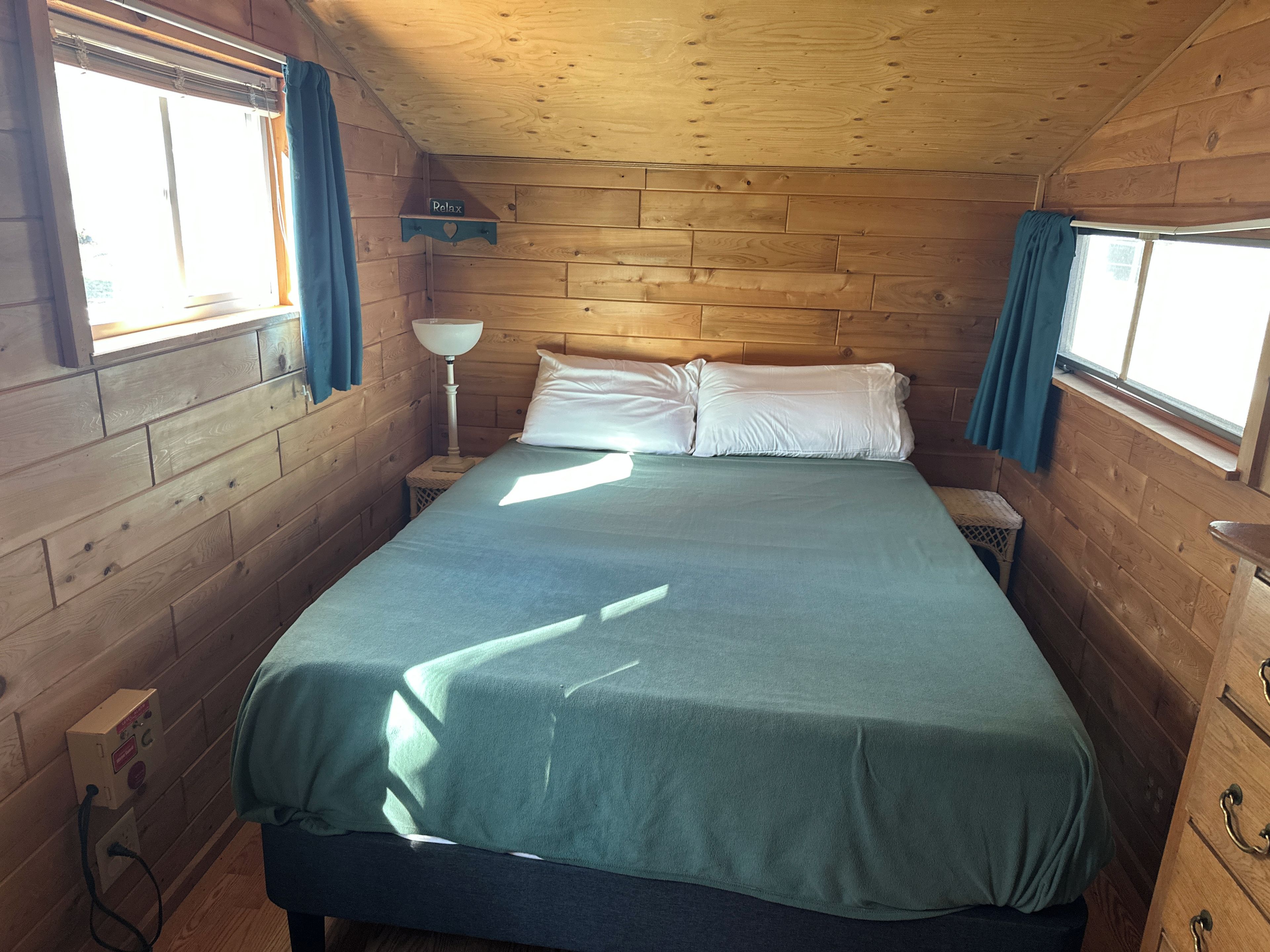 A wooden cabin room with a bed, a nightstand, a lamp, and a window with curtains.