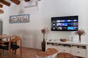 A living room with a flat-screen TV mounted on the wall, a wooden dining table with chairs, a white cabinet with drawers, and a decorative vase with dried flowers. - New Listing (Rab)