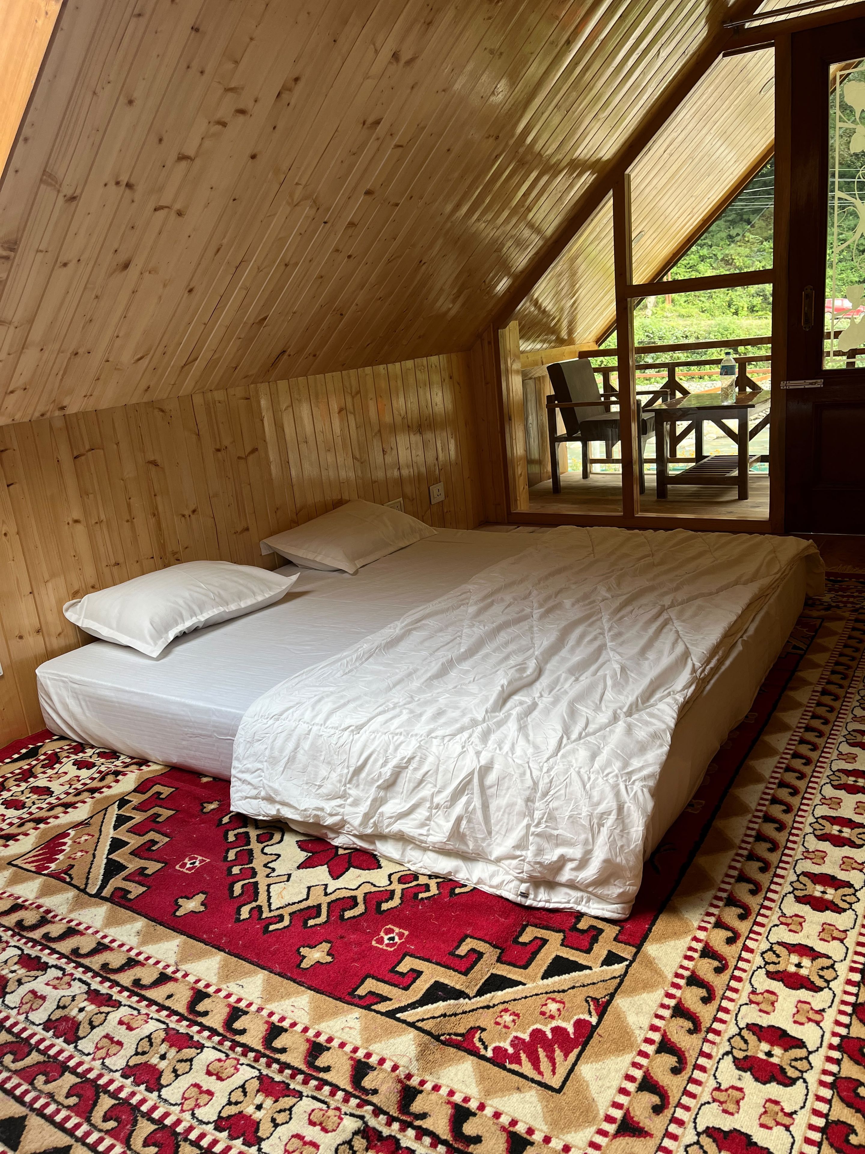 A bedroom with a wooden sloped ceiling, a bed with white bedding, a red patterned rug, and a view of an outdoor patio with chairs and a table.
