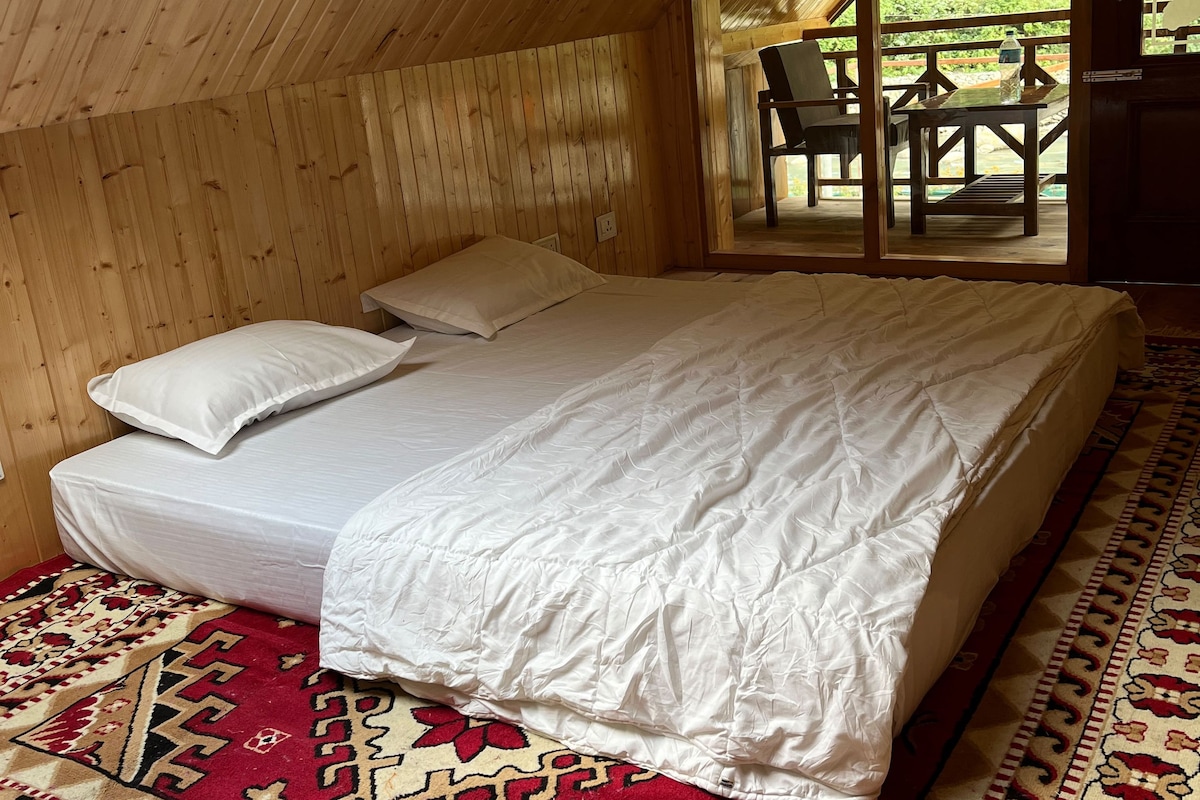 A bedroom with a wooden sloped ceiling, a bed with white bedding, a red patterned rug, and a view of an outdoor patio with chairs and a table.