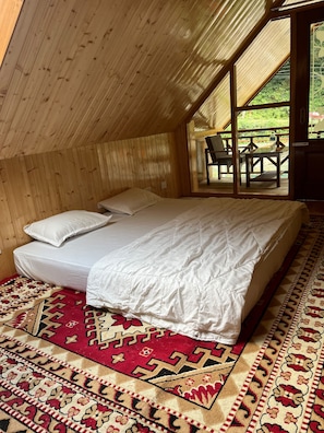 A bedroom with a wooden sloped ceiling, a bed with white bedding, a red patterned rug, and a view of an outdoor patio with chairs and a table. - Himalayan River Nook Cottages (Sainj)