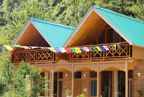 A building with a wooden facade and a green roof, featuring a balcony with colorful flags. - Himalayan River Nook Cottages (Sainj)
