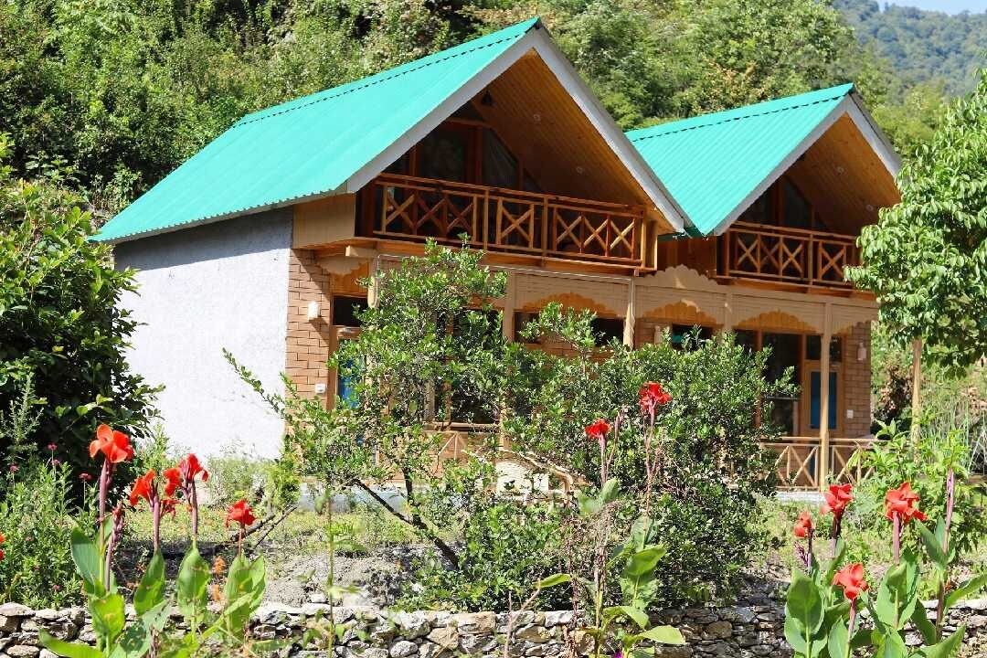 Una cabaña de madera con techo verde, rodeada de un jardín con flores rojas.