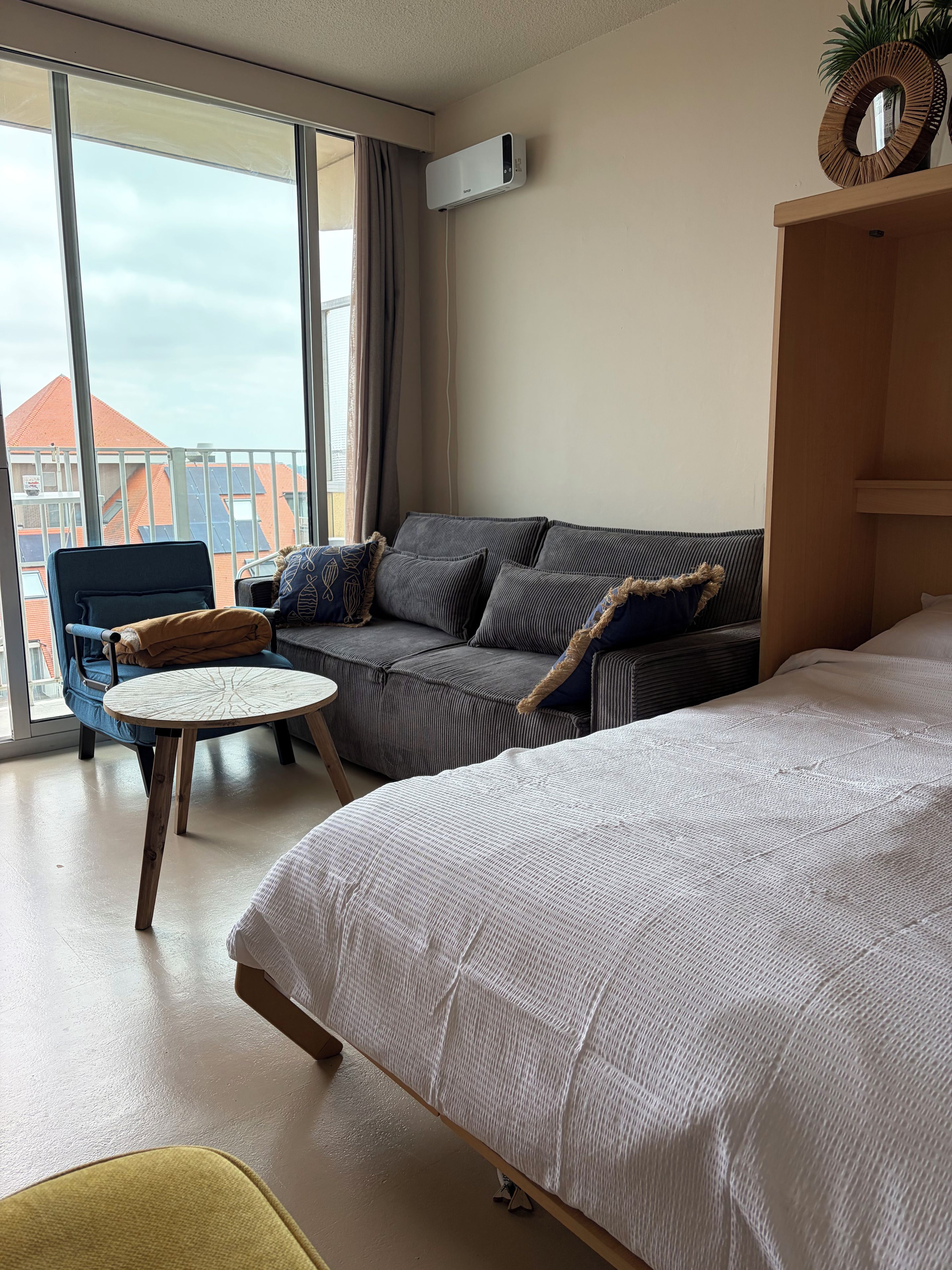 Une chambre d’hôtel avec un lit, un canapé, une petite table ronde et une chaise. Il y a une fenêtre avec des rideaux, et on peut voir une plante sur le rebord de la fenêtre.
