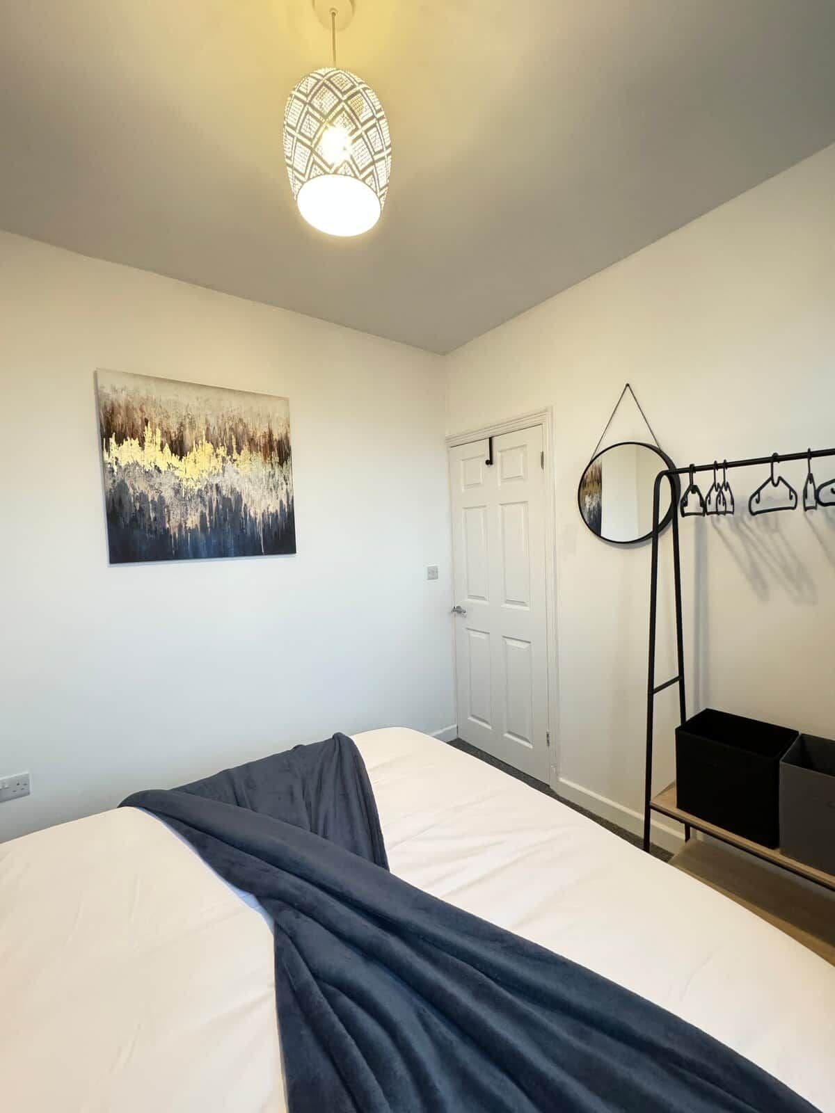 Un dormitorio con cama, lámpara colgante, puerta, perchero y un cuadro en la pared.