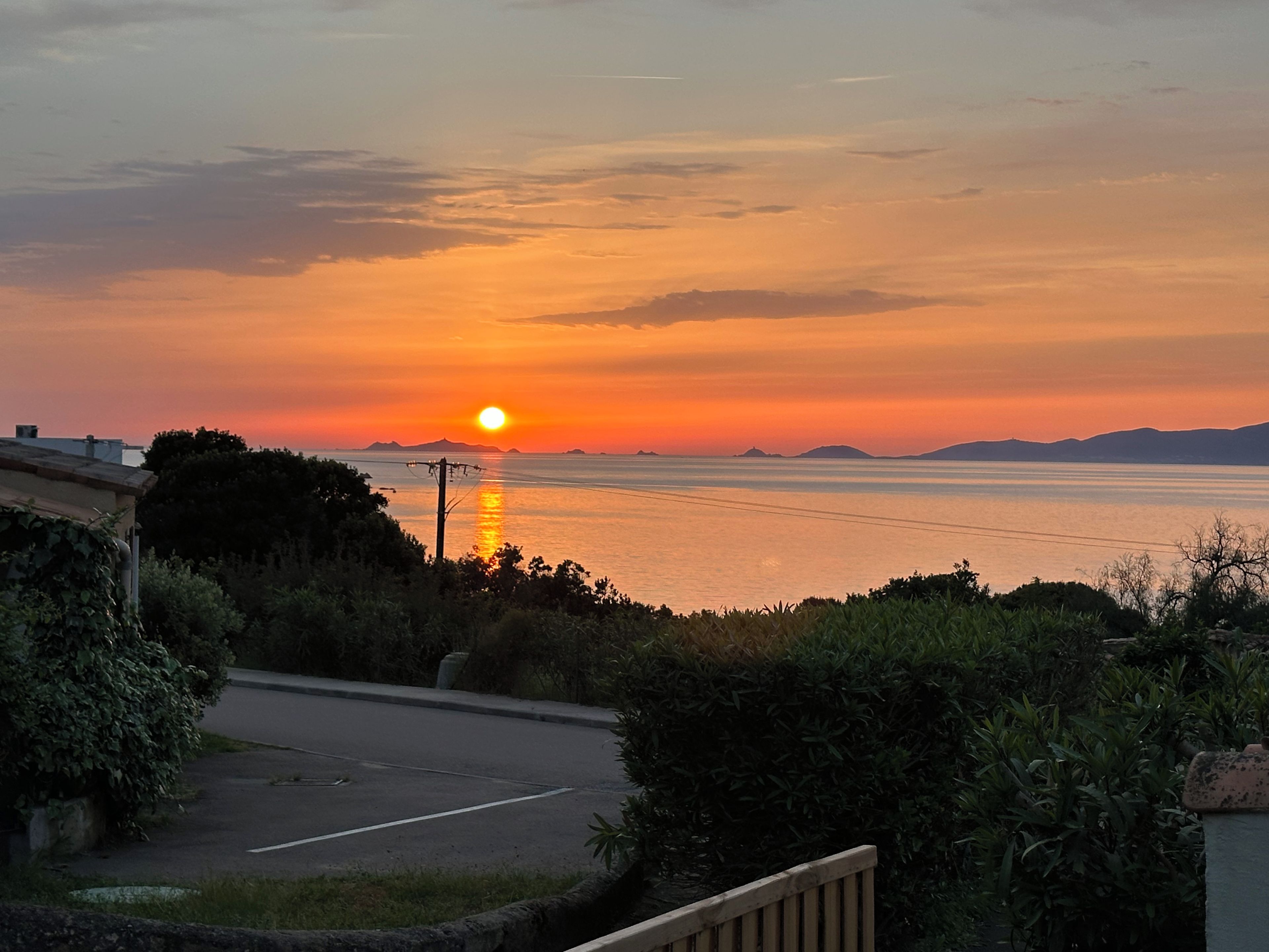 A sunset over a body of water with distant landforms, a road, and vegetation in the foreground.
