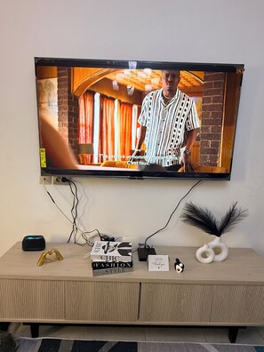 A television mounted on a wall with a flat-screen display. Below the TV, there is a wooden console with a few items on it, including a book titled 'The Fashion in Style,' a small decorative item, and a black and white vase. - 2-bedroom apartment in enchanting Douala with AC comfort and luxury (Douala)