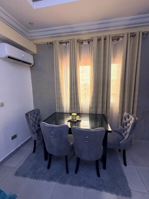 A dining area with a glass table surrounded by four chairs, a small vase, and a window with curtains. - 2-bedroom apartment in enchanting Douala with AC comfort and luxury (Douala)