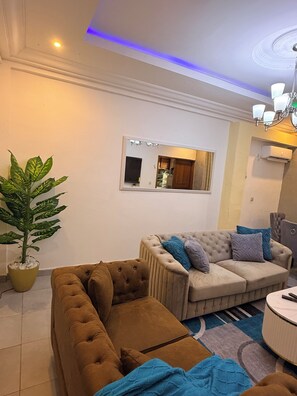 A living room with a brown sofa, a beige sofa with blue cushions, a potted plant, and a mirror on the wall. - 2-bedroom apartment in enchanting Douala with AC comfort and luxury (Douala)